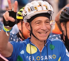 Smiling man with white bike helmet and blue shirt holding right hand up making V with fingers