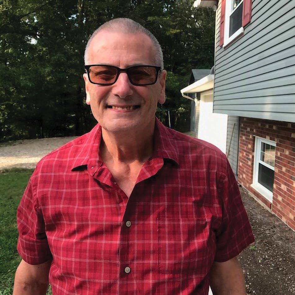 Garry Still, wearing a short-sleeved red plaid button up shirt, smiling, with his house in the background
