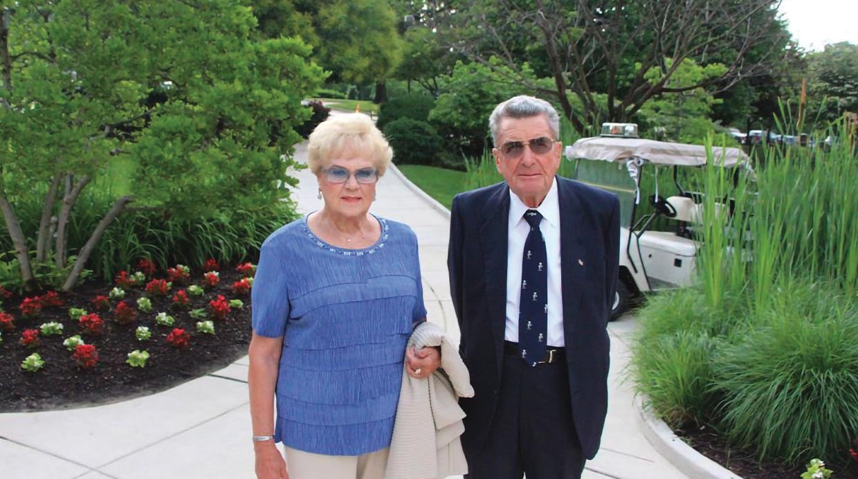 Guadalupe and Albert Folgueras standing in a pathway. There is a golf cart in the background and landscaping with trees, grass and flowers.