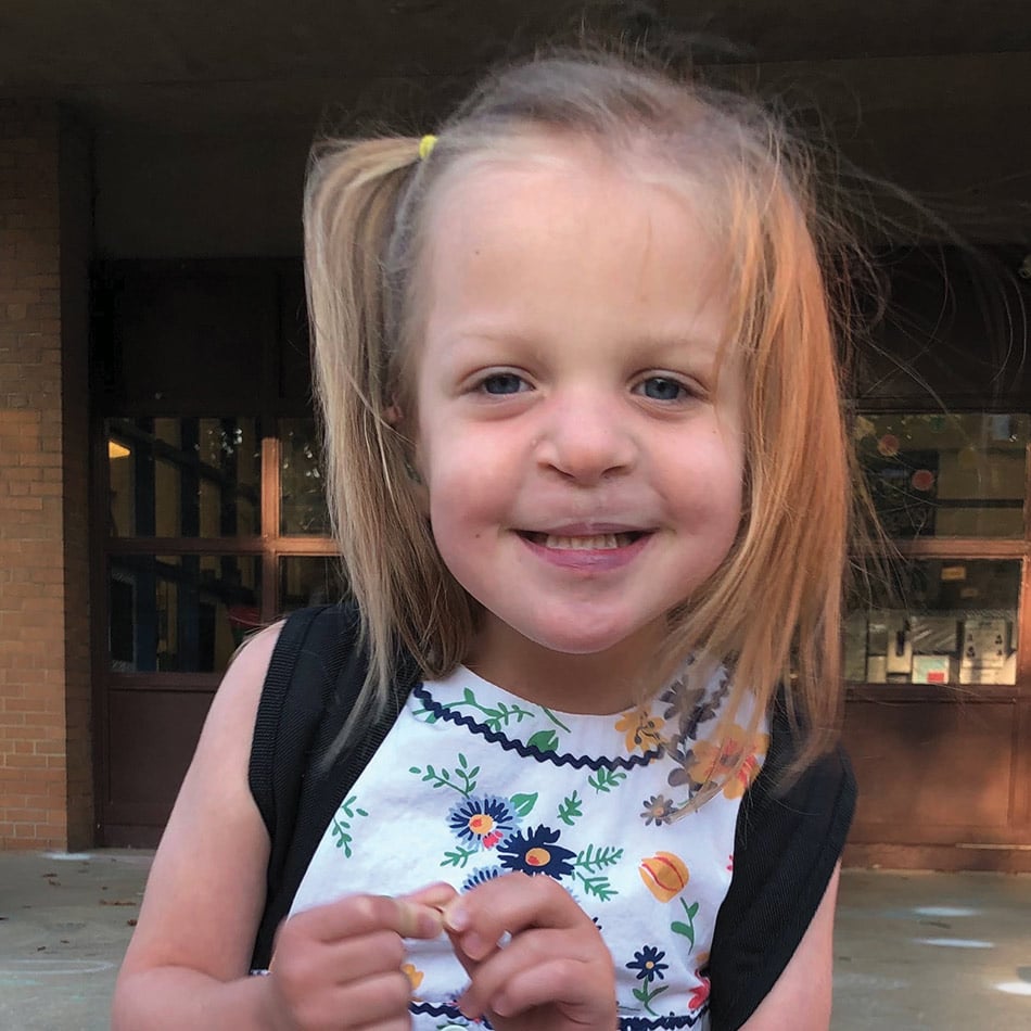 Charlotte, with blonde hair and a side half pony tail, smiling for a photo in front of her preschool
