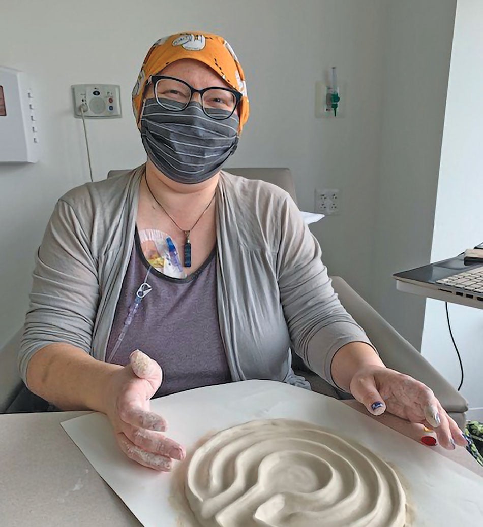 Lydia sitting at a table with clay on the surface. She is wearing a face-mask but is smiling underneath. She is also wearing a head scarf and glasses