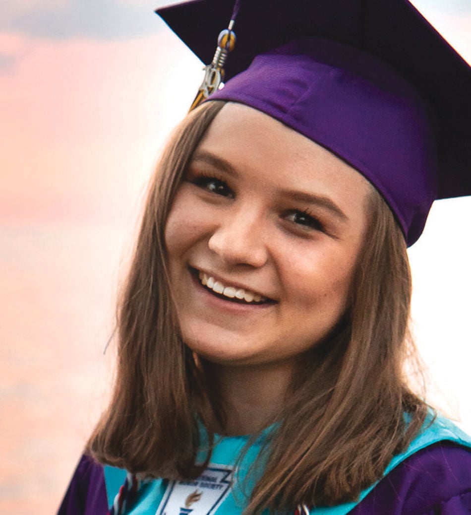 Erin Black, a young woman, wearing a purple cap and gown, smiling at the camera in a graduation photo, with a pink sunset behind her