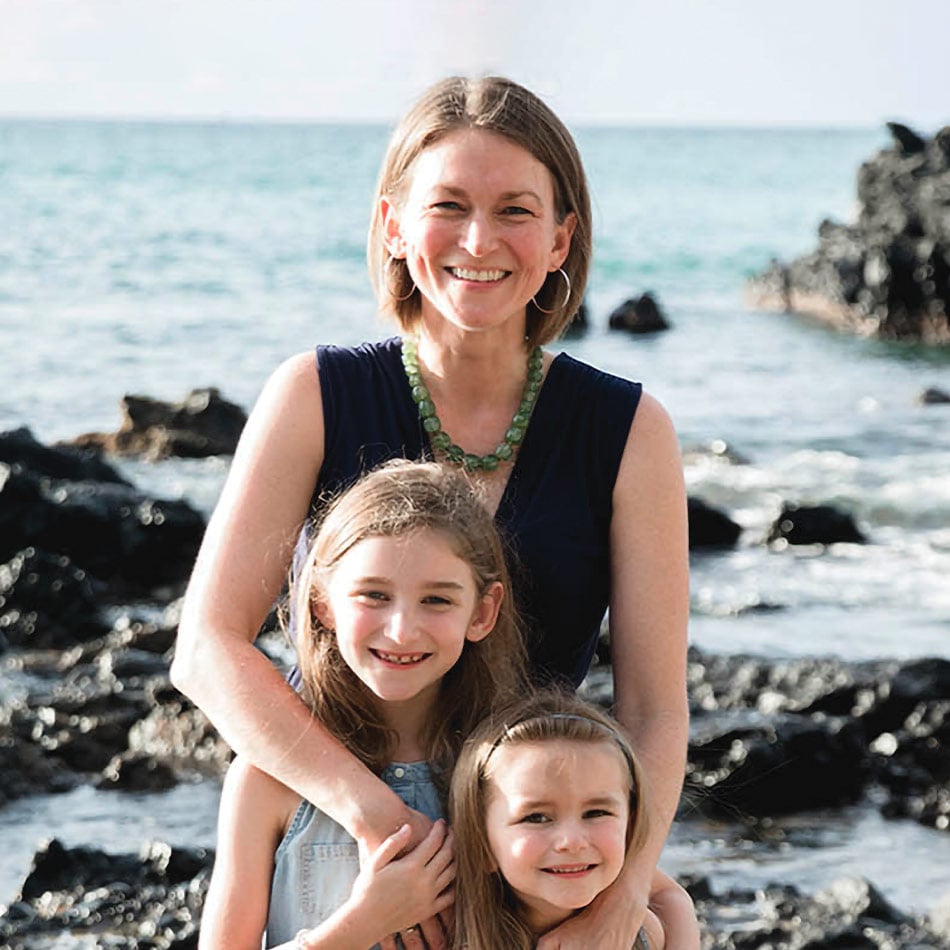 Kate Watson and her two daughters, standing in front of a beach with water and rocks behind her