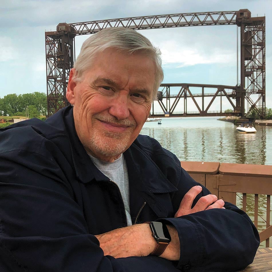 Rick Holmes sitting at a table with this arms crossed and smiling at the camera. Behind him is the Cuyahoga River with a bridge and a boat in the water