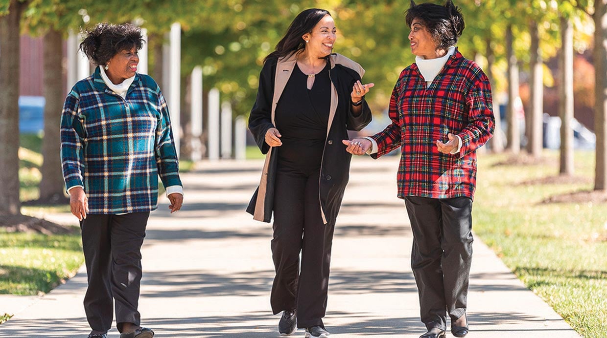 Three women, walking on a paved path talking about smiling.