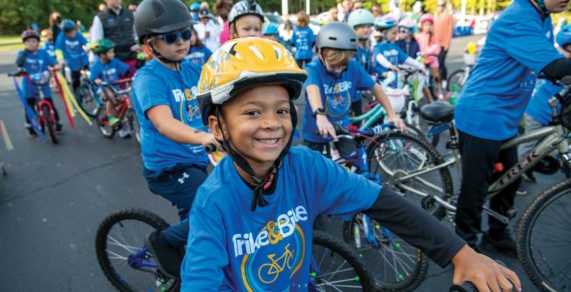 Crowd at bicycle race focused on black boy with yellow helmet wearing blue shirt that says Trike & Bike