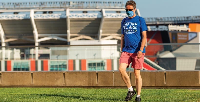 Man walking on grass behind stadium wearing sunglasses medical mask blue Velosano shirt red shorts white socks black shoes