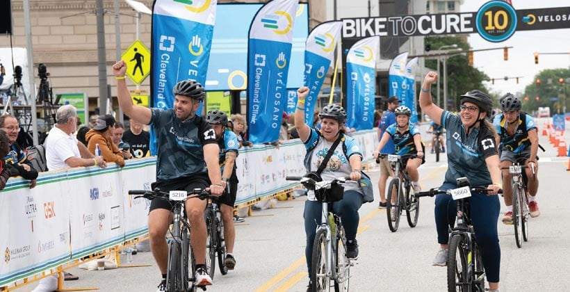 People finishing bicycle race with right hand raised in fist in front of signs Bike to Cure Velosano Cleveland Clinic