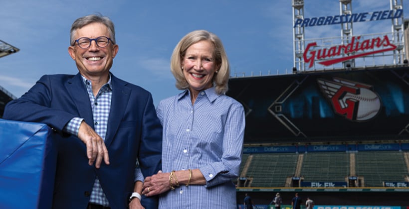 Smiling couple man glasses blue blazer blue and white plaid shirt woman blue gingham longsleeved shirt standing in front of Cleveland Guardians baseball stadium 