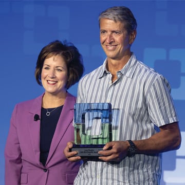 Woman in purple blazer next to man in gray striped shirt holding award
