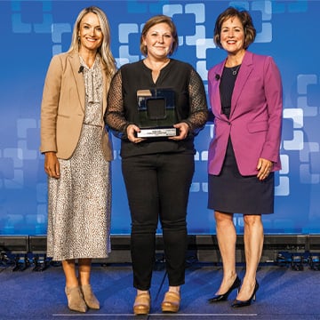 Three woman on a stage with blue patterned backdrop center woman holding award