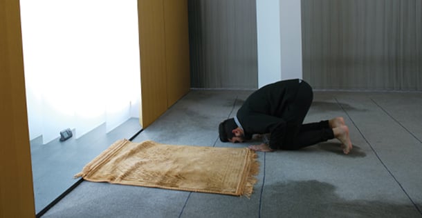 Man kneels with his face to the floor next to a blanket in front of a window to pray
