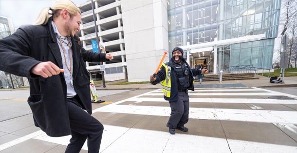 man with blond ponytail black overcoat gray shirt black jeans hightops dancing on sidewalk next to smiling black man crossing guard