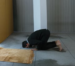 Man kneels with his face to the floor next to a blanket in front of a window to pray