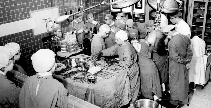 Black and white photo of 1950s operating room with a dozen doctors and nurses performing surgery