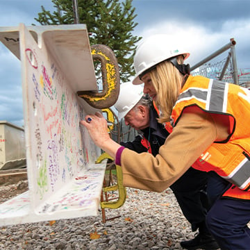 Man and woman in hard hats and orange vests squat to sign I-beam at construction site