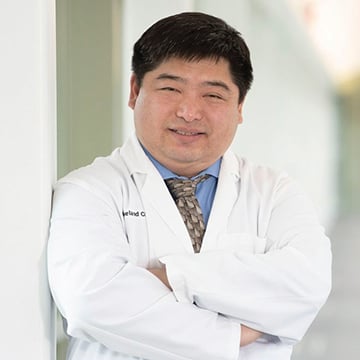 Smiling Asian doctor in lab coat arms crossed leaning against wall