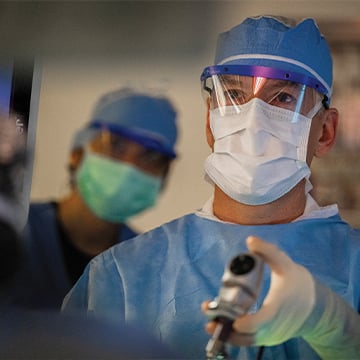Doctor wearing surgical scrubs mask gloves holding tool looking at screen in surgery
