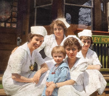 Four female nurses wearing white uniforms with a small child in the middle, looking at the camera