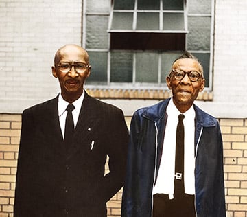 two older man of color, standing side by side. one wearing a suit and tie, the other where a leather jacket and tie. 