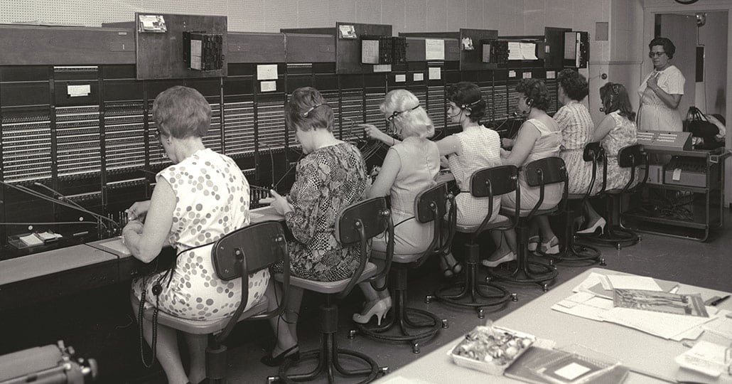 8 women, 7 who are sitting at the phone operator's table, connecting lines