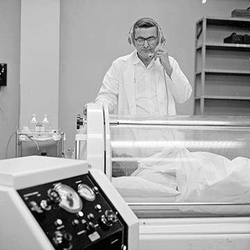 Dr. Wasmuth at a hyperbaric oxygen chamber with a patient inside, speaking into a headset. Centennial-ThinkingBig-Floated