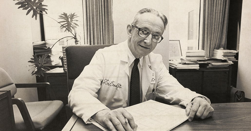 William Proudfit, MD., sitting at this desk with paper on the tabletop, looking at the camera
