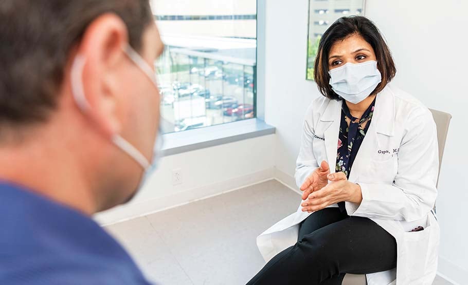 Female doctor in white lab coat, seated, talking to a man. both wearing face masks.