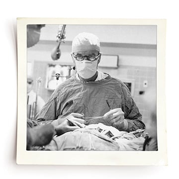 Black and white photo of doctor in an operating room holding medical tools. He is wearing a scrub cap, face mask, glasses and a surgical cape.