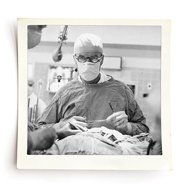 Black and white photo of doctor in an operating room holding medical tools. He is wearing a scrub cap, face mask, glasses and a surgical cape. 