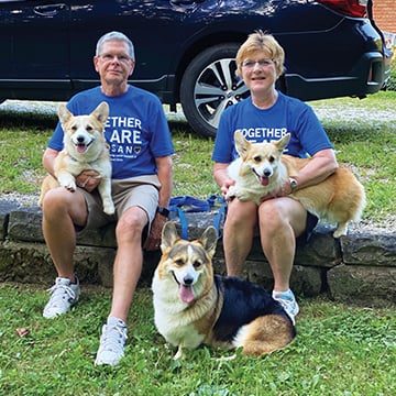 A man and a woman sitting outside, both holding a corgi, and another corgi is sitting in the grass in between them