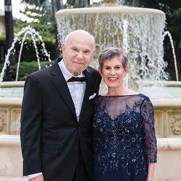 Sandy (right) Fox, DDS standing with an older man, wearing formal attire, in front of an outdoor fountain.