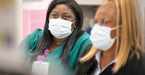 Aryanna Lewis sits in green scrubs and a mask on the left, Evelyn Penn sits in a black suit jacket and mask on the right.