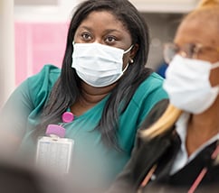 Aryanna Lewis sits in green scrubs and a mask on the left, Evelyn Penn sits in a black suit jacket and mask on the right.