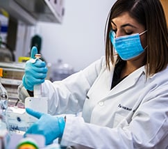 Female lab technician wearing a mask, gloves, and white lab coat administers a sample in a medical lab.
