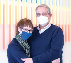 Cathy and Kevin Poulsen wearing face masks and navy blue sweaters, embracing and looking at the camera in front of a art wall.