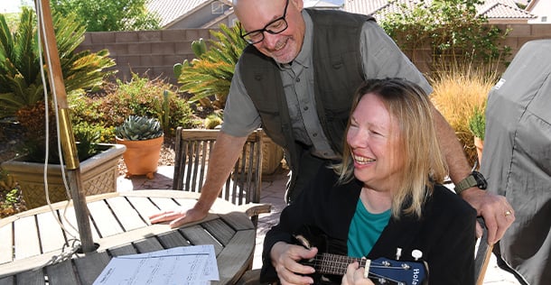 A woman plays guitar on the left while a man leans over her outside at a table on a patio.