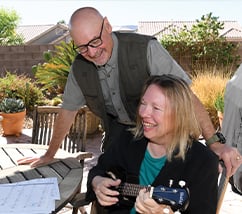 A woman plays guitar on the left while a man leans over her outside at a table on a patio.