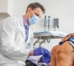 Doctor wearing mask and white coat leans over a patient's knees to administer an injection in a white clinic room.