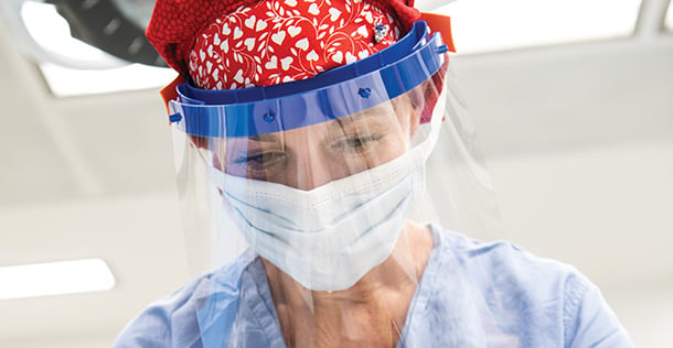 Woman wearing light blue scrubs, a red scrub cap with white hearts, surgical mask, and face shield