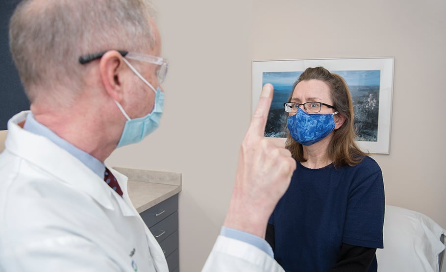 A male doctor wearing a surgical mask, waving his finger in the air while the patient across from him looks at it.