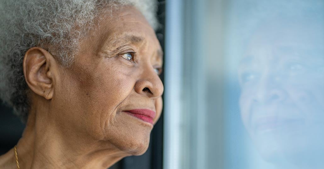 Older woman looking out of a window, her reflection appears in the glass of the window