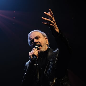 Neil Diamond wearing a black shiny blazer, raising his left hand to the sky while holding a microphone in the other, singing