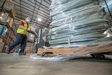 A male worker wearing a safety vest and mask, pushing a pallet lift of medical supplies in a warehouse