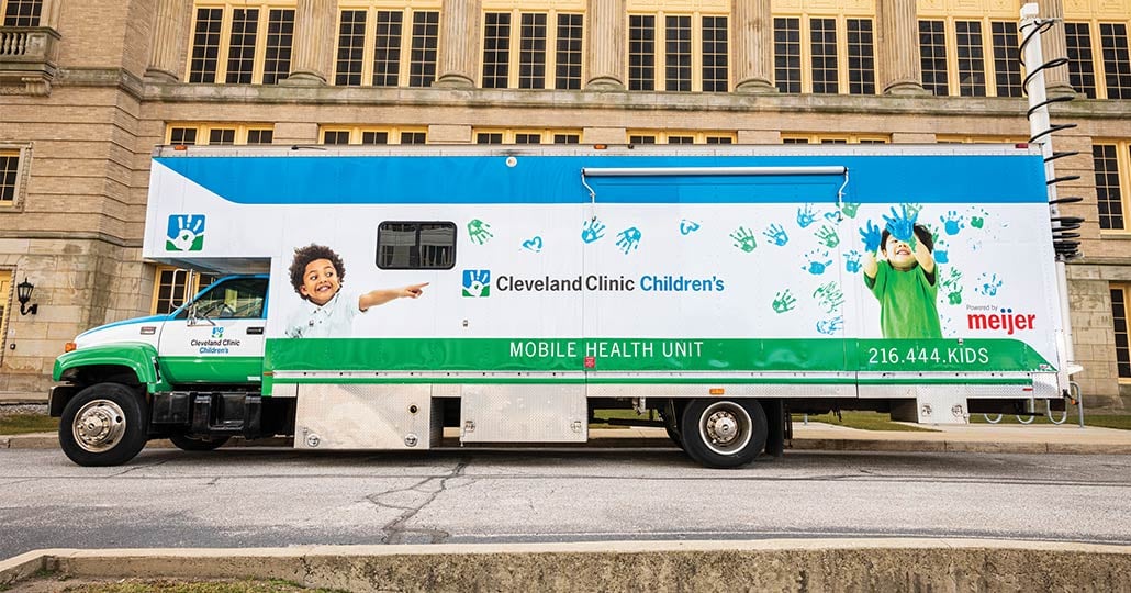 A shipping truck parked outside of a beige building. The truck as graphic design on the side that shows images of kids playing with paint. The graphics says 