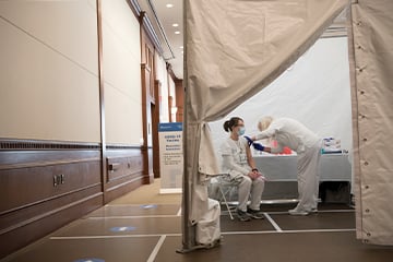 Two nurses in a white tent. One administering a shot into the other nurse who is sitting in a chair. 