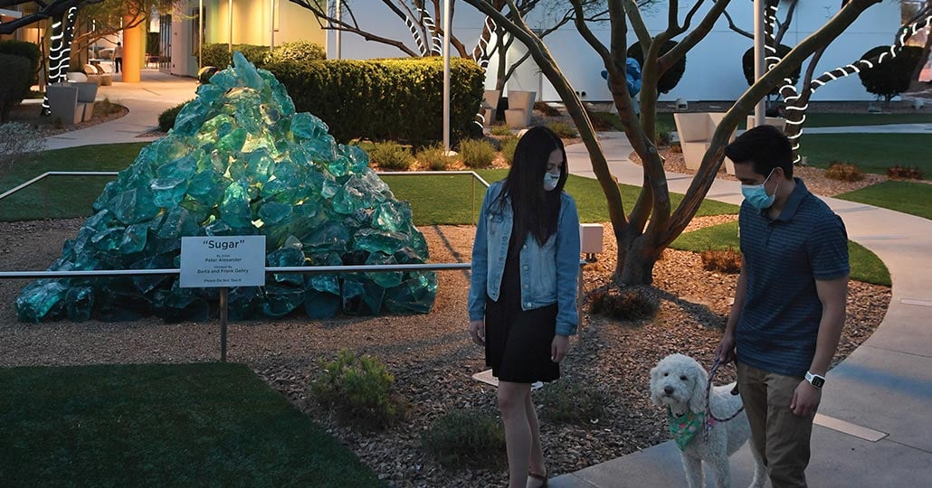 A man and a woman walking a dog in a park with a turquoise glass pyramid sculpture in the background