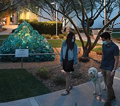 A man and a woman walking a dog in a park with a turquoise glass pyramid sculpture in the background