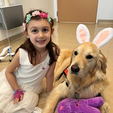 Girl in white dress with colorful flower headband sitting next to golden retriever wearing bunny ears headband