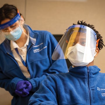 a person wearing PPE masks and a blue zipped jacket sitting in front of a person standing wearing PPE masks and a blue zipped jacket